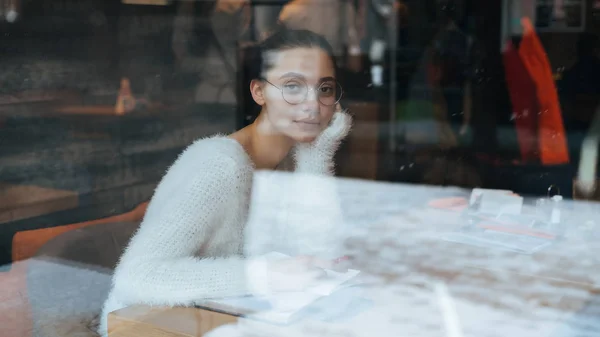 Мила дівчина в білій куртці і окулярах сидить в кафе, відпочиває після навчання — стокове фото