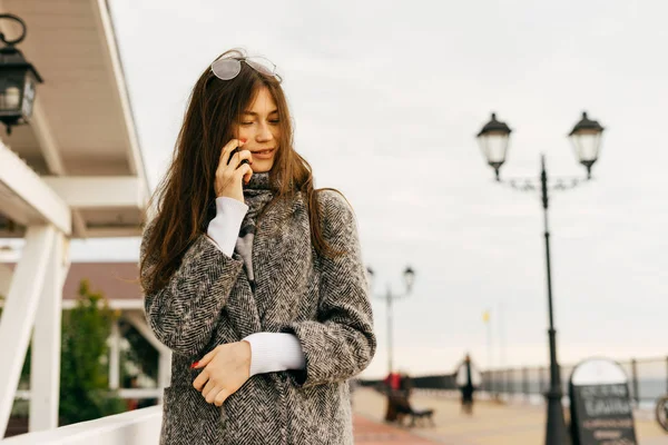 Jeune fille brune en manteau gris à la mode marchant à l'extérieur, parlant au téléphone, style de rue — Photo