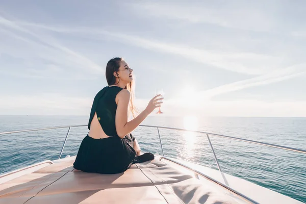 Роскошная молодая женщина в черной летней одежде сидит на своей белой яхте, пьет шампанское и наслаждается отдыхом — стоковое фото