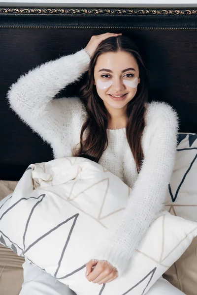 Bela menina de cabelos longos em pijama branco senta-se na cama, no início da manhã, sob os olhos de manchas brancas, sorrindo — Fotografia de Stock