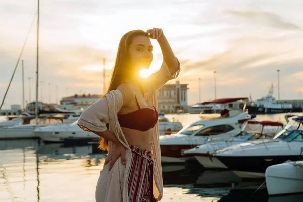 Lussuosa ragazza dai capelli lunghi in eleganti vestiti estivi in posa nel porto marittimo ai raggi del sole della sera — Foto Stock