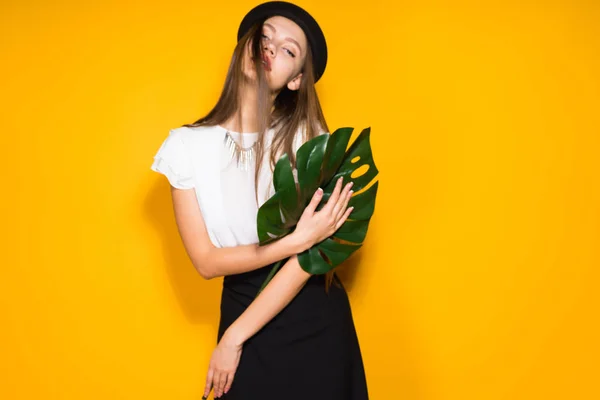 Atractiva chica de pelo largo modelo en sombrero negro sostiene una hoja verde, posando sobre fondo naranja — Foto de Stock