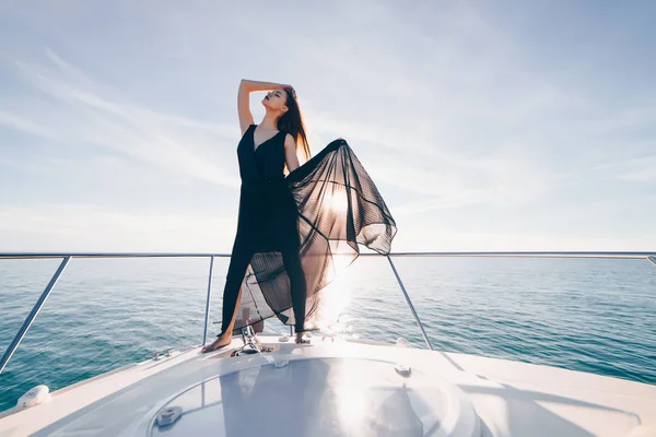 Молодая красивая девушка в черной летней одежде стоит на своей белой яхте и позирует на солнце — стоковое фото
