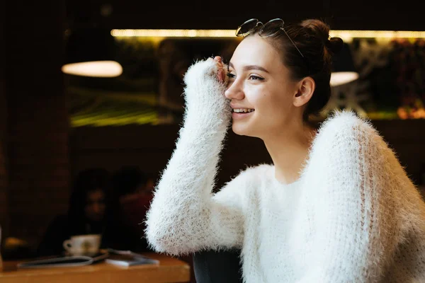 Милий молода дівчина студент в білій куртці сидить в кафе після навчання в університеті, дивлячись у вікно і посміхаючись — стокове фото