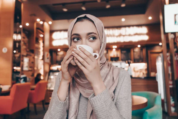 Красивая арабская девушка с платком сидит в уютном восточном кафе, пьет кофе и отдыхает — стоковое фото