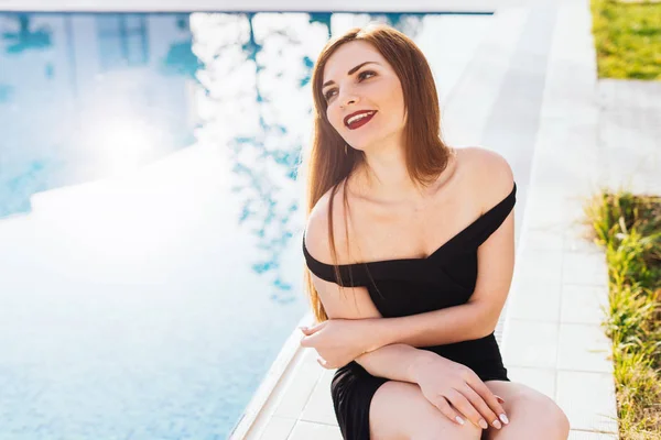 happy rich woman in a black dress sitting in her garden by the pool in the sun