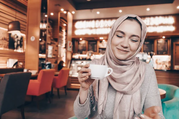 Belle fille arabe avec un foulard assis dans un café confortable, boire du café et rire — Photo