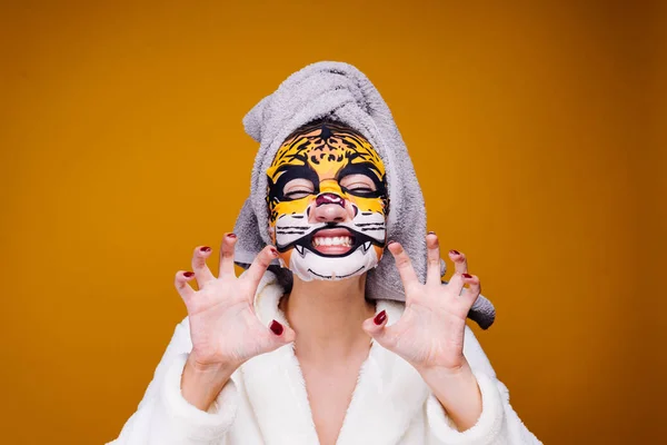 Una joven divertida con una toalla en la cabeza y en un albornoz ruge como una bestia, una máscara con una cara de leopardo en su cara — Foto de Stock