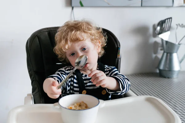 Кудрявый маленький мальчик сидит в детском кресле на кухне, ест здоровую кашу — стоковое фото