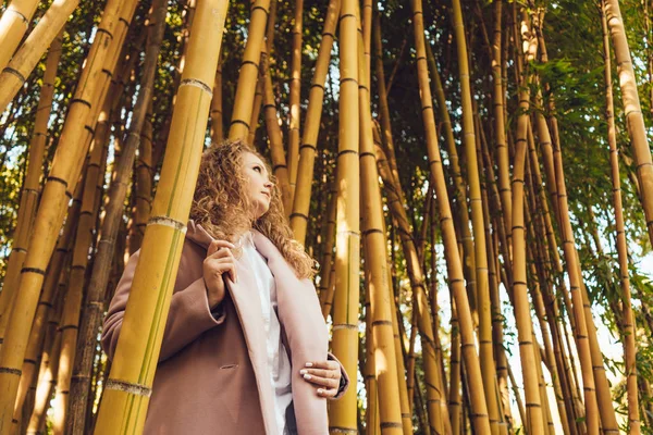 Mujer joven y rizada disfrutando del clima primaveral, parada junto a altos árboles de bambú — Foto de Stock