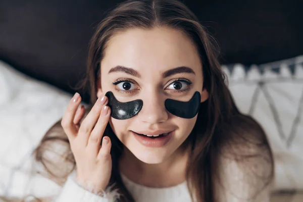 Carino ragazza dai capelli lunghi in pigiama bianco seduto sul letto la mattina presto, sotto gli occhi di macchie nere, sorridente — Foto Stock