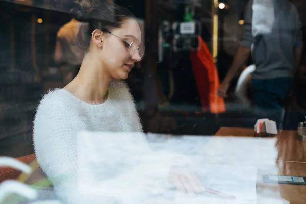 Задумчивая молодая девушка-фрилансер в белой куртке и очках сидит в кафе и думает о работе — стоковое фото