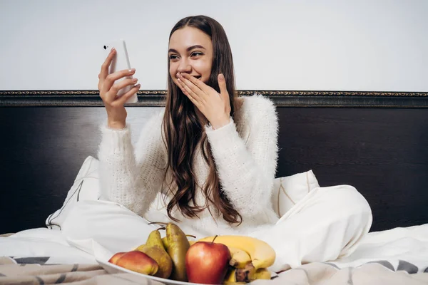 Una linda chica de pelo largo en pijama blanco se sienta en la cama temprano en la mañana, desayunos con frutas útiles, mira en el teléfono inteligente y se ríe — Foto de Stock