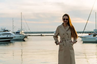 liman içinde akşam poz ve gülümseyen bir moda bej bir palto şık uzun saçlı kız