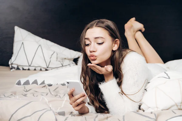 Encantadora joven se acuesta en la cama, descansando, hace selfie y envía un beso de aire — Foto de Stock