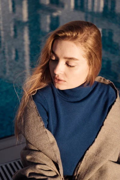 Mooie jong blond meisje in blauwe trui zit bij het zwembad buiten in de zon — Stockfoto