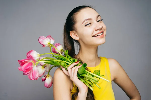 Sarı elbiseli mutlu genç kız pembe çiçekler tutar, gülüyor ve kameraya benziyor — Stok fotoğraf