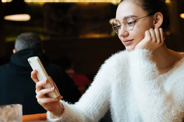Cute młoda dziewczyna student siedząc w kawiarni po studiach na Uniwersytecie, w okularach patrząc na smartphone — Zdjęcie stockowe
