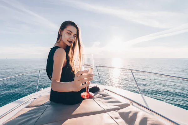 Красивая роскошная девушка в черном платье сидит на своей белой яхте, пьет шампанское и наслаждается богатой жизнью — стоковое фото