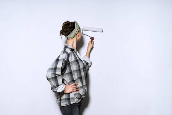 молодая девушка в клетчатой рубашке красит стены платформой, делает ремонт в своей новой квартире
