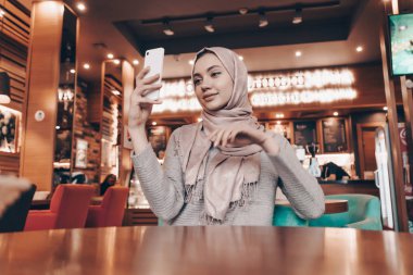 Ona hijab bir başörtüsü bir Arap kızı rahat bir kafede oturur ve onu Smartphone'da selfie yapar