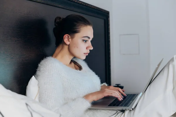 Chica joven enfocada en pijamas blancos sentada en la cama, trabajando en el portátil — Foto de Stock