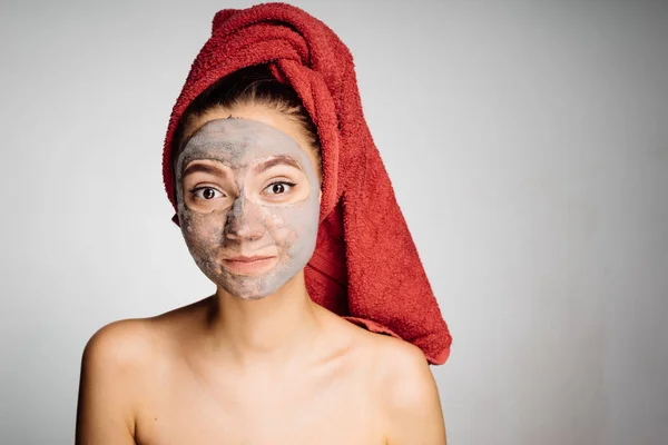 En söt ung flicka med en röd handduk på huvudet tillämpas en användbar lermask till ansiktet, dagen av spa — Stockfoto