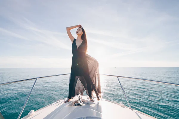 Стильная молодая девушка в черном летнем платье стоит на своей белой яхте, наслаждается морским вояжем — стоковое фото