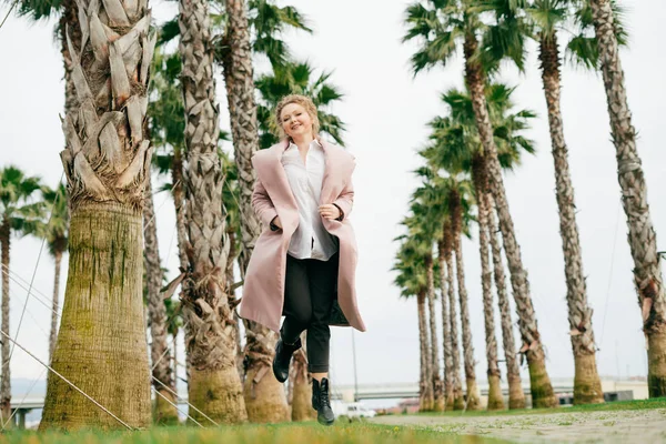 Happy young woman in a stylish coat walking through a green park, enjoying spring and tall palms — Stock Photo, Image
