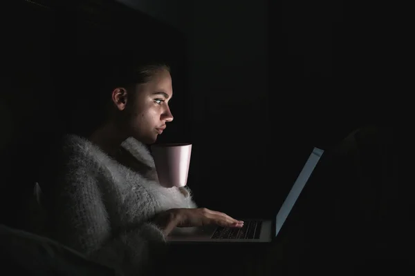 Зацікавлена молода дівчина сидить у ліжку в темряві, дивиться фільм на ноутбуці і п'є чай — стокове фото
