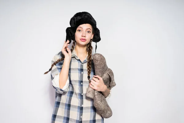 Piękny rosyjski dziewczyna z warkoczykami, w ciepłe futro kapelusz posiada szare zimowe buty i patrzy w kamerę — Zdjęcie stockowe