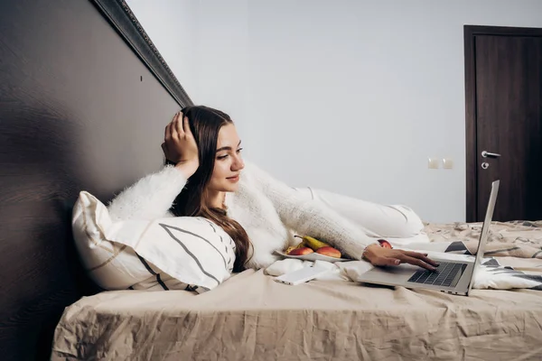 Een jong meisje in witte pyjama ligt op een bed, rust na een dag werken, kijken naar een film op haar laptop — Stockfoto