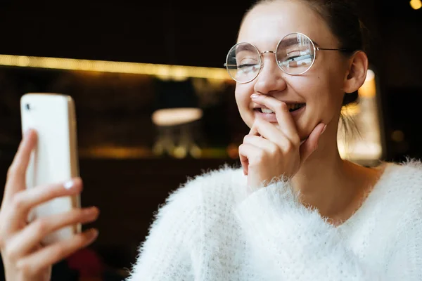 Freelancer piękna młoda dziewczyna w okularach, siedząc w kawiarni, patrząc na swoim smartfonie i śmiejąc się — Zdjęcie stockowe