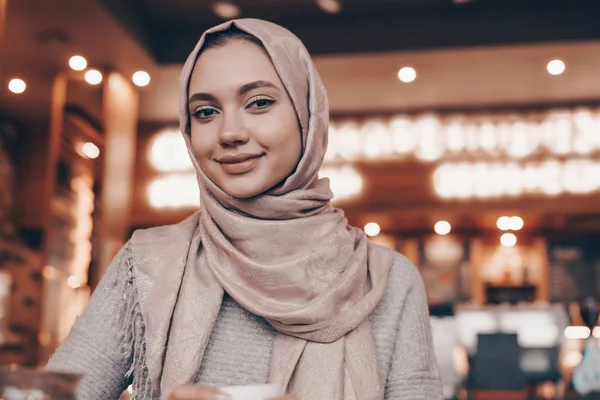 Linda chica árabe con un pañuelo en la cabeza sentado en un acogedor café oriental, sonriendo y mirando a la cámara — Foto de Stock