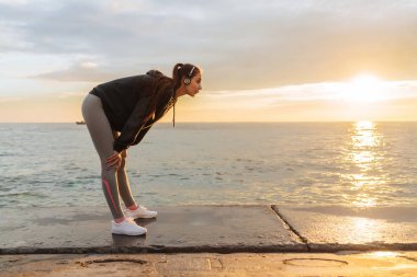 dinlenmek için ince atletik kız ishal günbatımı, kulaklıklar, müzik dinler, deniz kenarında durdu