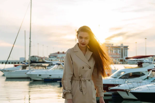 Lussuosa ragazza dai capelli lunghi in un elegante cappotto beige in posa nel porto marittimo ai raggi del sole della sera — Foto Stock