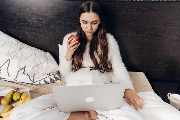Серйозна довгошерста дівчина в піжамі лежить в ліжку, виглядає цікаво епізод на ноутбуці і їсть яблуко — стокове фото