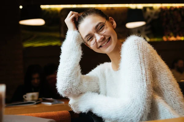 Мила молода дівчина студент в окулярах і біла куртка сидить в кафе, дивлячись на камеру і сміючись — стокове фото