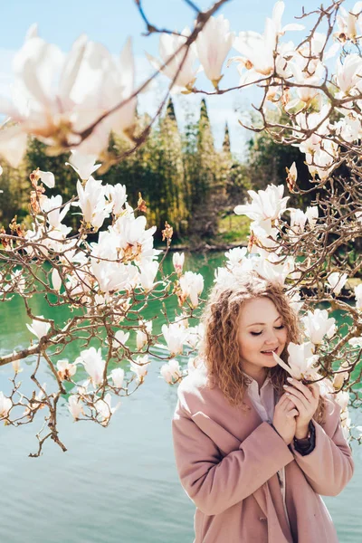 Joven rizada en un abrigo rosa huele una magnolia floreciente aromática, en un parque al lado de un estanque, sonríe — Foto de Stock