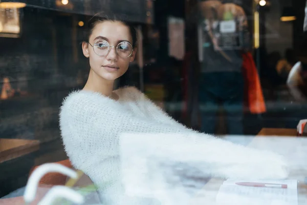 Молода красива студентка в окулярах сидить в кафе після навчання в університеті і дивиться у вікно — стокове фото