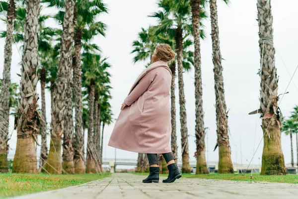 Élégante jeune femme dans un manteau rose se promène dans le parc à côté de grands palmiers verts, le printemps et la chaleur — Photo