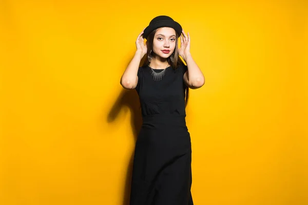 Hermosa chica de moda en vestido elegante negro y sombrero posando sobre fondo naranja, modelo — Foto de Stock