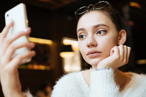 Piękna młoda dziewczyna student w białą kurtkę jest siedząc w kawiarni i patrząc na jej smartphone, myśląc o studiach — Zdjęcie stockowe