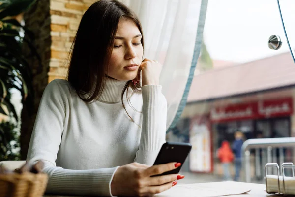 Pensive dark-haired girl sitting in cafe after a walk, looking into her smartphone — Stock Photo, Image