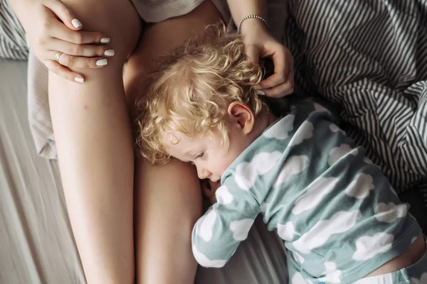 Pequeño bebé rizado abraza a mamá por la pierna, se acuesta en la cama y descansar — Foto de Stock