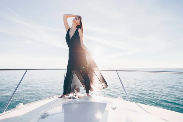 A luxurious rich girl in a black summer dress stands on her white yacht, enjoys sea voyage — Stock Photo, Image