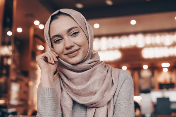 Hermosa joven árabe con pañuelo en la cabeza sonriendo y mirando a la cámara, sentado en la cafetería — Foto de Stock