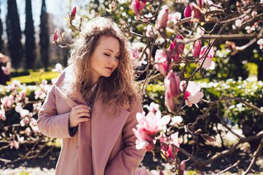 kıvırcık saçlı kız pembe bir ceket sıcak bahar gününde parkta yürüyor, güzel kokulu bir manolya sahiptir