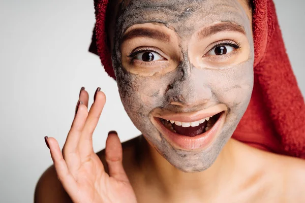 Счастливая молодая девушка с красным полотенцем на голове надела полезную глиняную маску на лицо, улыбаясь — стоковое фото