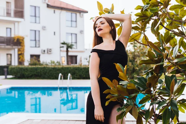 Uma mulher rica de luxo em um vestido preto andando através de seu jardim junto à piscina azul — Fotografia de Stock